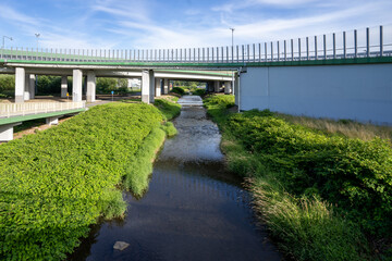 Rzeka płynąca przez tereny przemysłowe, w dali zjazd z autostrady na betonowych palach.