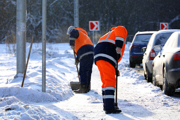 Two men workers in uniform with a shovels cleaning the street near the car parking. Snow removal in winter city at frost weather