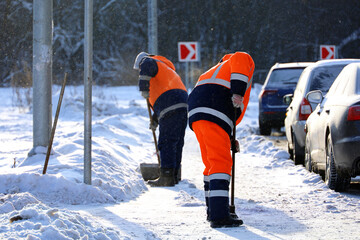 Two men workers in uniform with a shovels cleaning the street near the car parking. Snow removal in...