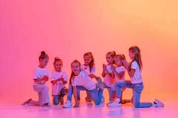 Happy kids, school age girls in white t-shirts and jeans dancing in choreography class isolated on pink background in yellow neon light.