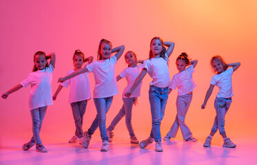 Group of children, school age girls in sportive casual style clothes dancing in choreography class isolated on pink background in yellow neon light.