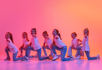 Naklejka premium Happy kids, school age girls in white t-shirts and jeans dancing in choreography class isolated on pink background in yellow neon light.