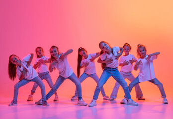 Group of school age girls, cheerful kids in casual style clothes dancing, jumping isolated on pink background in yellow neon light. Music, dance, art