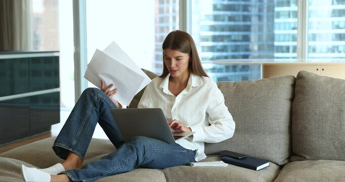 Young attractive woman sit on cozy sofa in modern living room use laptop, sorts out papers, manage family household, pay bills on-line. Student female prepare for exams at home, studying looks focused