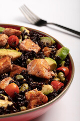 A red bowl of black rice with baked salmon, avocado, tomatoes, chili peppers and edamame.
