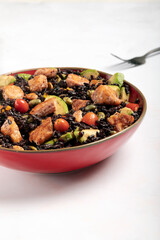 A red bowl of black rice with baked salmon, avocado, tomatoes, chili peppers and edamame.