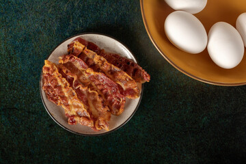 Crispy bacon slices in a white little dish beside some fresh eggs.