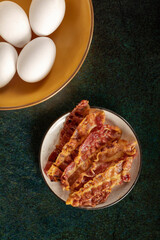 Crispy bacon slices in a white little dish beside some fresh eggs.