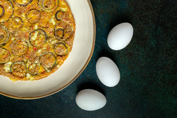 Homemade onion omelette in a white dish beside three fresh eggs on a raw dark background.