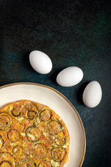 Homemade onion omelette in a white dish beside three fresh eggs on a raw dark background.