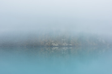 Simple meditative landscape with turquoise lake in thick fog. Tranquil view through clearance in...