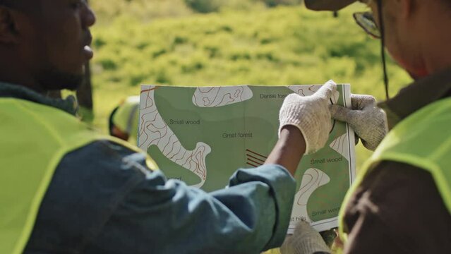 Over-shoulder of two male cropped geographers holding map of area, standing in forest on sunny spring day, talking