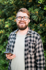 Fototapeta premium Portrait of attractive cheerful guy using smartphone for scrolling on social media over green tree background