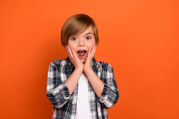 Photo of young boy hands touch cheeks astonished speechless novelty isolated over orange color background