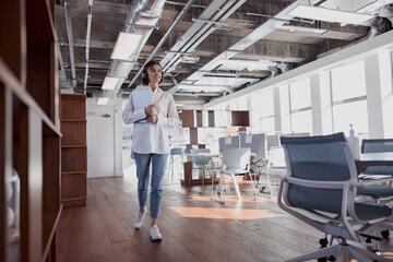 Fototapeta premium Attractive business woman in headphones with laptop crossing modern office. High quality photo