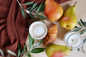 Cup of coffee with croissant, pears, candle, green branches on the light brown background. Fall season coffee. 