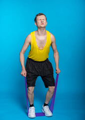 a guy in a yellow T-shirt and black shorts pulled on an elastic band, twisted his face. blue background. hard workout