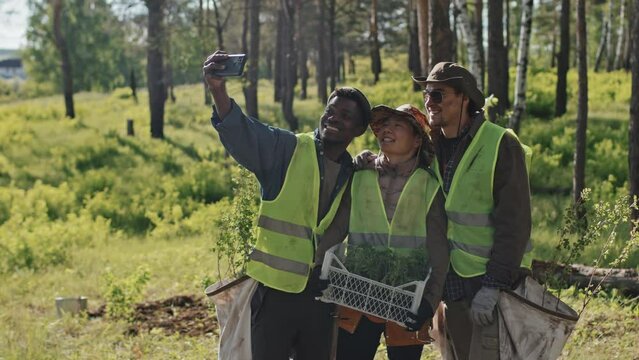 Medium long of three multiethnic eco volunteers with tree seedlings taking selfie in woods on sunny day