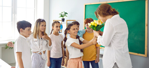 Fototapeta na wymiar Cute schoolchildren with flowers for their best female teacher at school in honor of holiday. Preteen classmates in classroom give teacher bouquet of tulips. Teacher's day concept. Web banner.