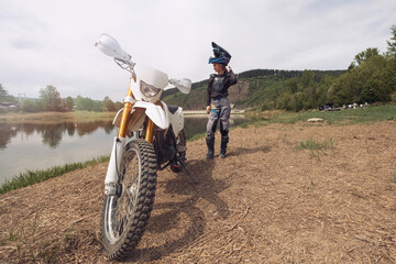 Biker female wearing moto protection and motocross helmet standing  with dirt motorcycle on river...