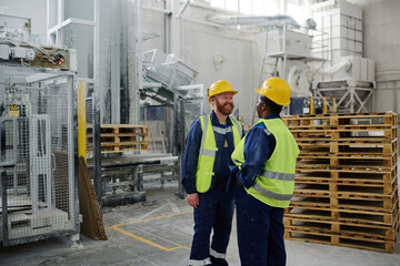 Cheerful bearded male engineer in uniform and safety helmet standing in front of female colleague during discussion of working points