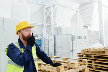 Young engineer of modern factory with walkie-talkie speaking to colleague while standing by stack of wooden pallets in workshop