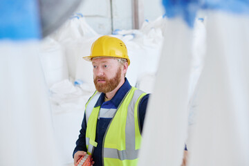 Bearded male worker in uniform and hardhat pressing buttons on remote control switch while lifting huge heavy sacks with raw materials