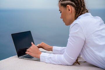 A woman is typing on a laptop keyboard on a terrace with a beautiful sea view. Close up of a woman's hands writing on a computer. Freelancing, digital nomad, travel and vacation concept.