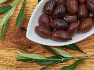 Kalamata olives in white bowl on olive wooden board with branch, top view