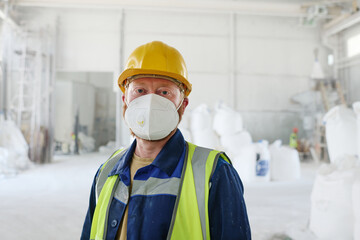 Worker of modern factory or distribution warehouse wearing protective respirator, workwear and safety helmet looking at camera