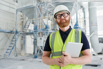 Confident bearded foreman in uniform and safety helmet holding tablet while standing in workshop of modern production factory