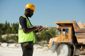 Side view of young black man in workwear and safety helmet using tablet while looking through online manual guide against truck