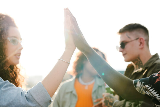 Young intercultural dates giving each other high five against their friends having chat during outdoor party in rooftop cafe or restaurant