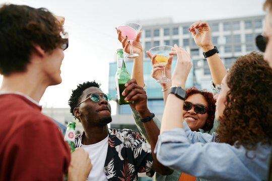 Group of cheerful friends raising hands with beer and cocktails while cheering up on terrace of rooftop cafe at outdoor party