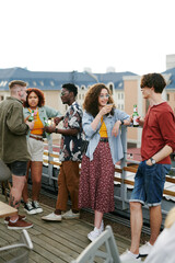 Young couple with drinks having chat against group of their friends with beer communicating at rooftop party on terrace of outdoor cafe