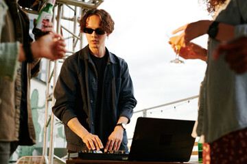 Young male deejay in casualwear and sunglasses making music for his friends with beer and cocktails enjoying rooftop party