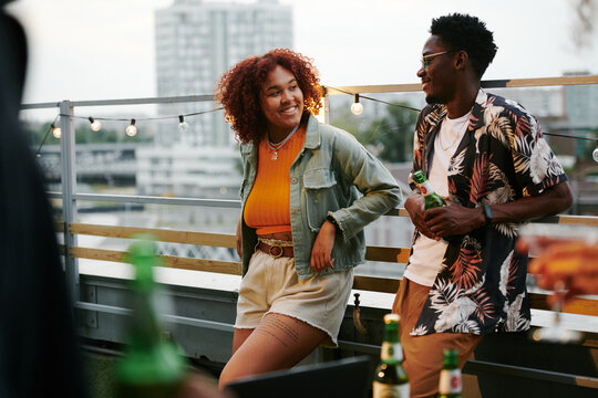 Young cheerful black woman in casualwear looking at her boyfriend during discussion of curious stuff on terrace of rooftop cafe