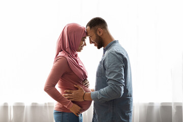 Pregnant Black Muslim Woman And Her Husband Hugging Near Window At Home
