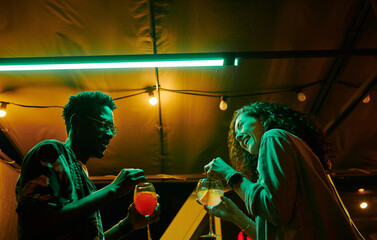 Happy young multicultural dates in casualwear having chat and cocktails while cheerful girl laughing during talk with boyfriend