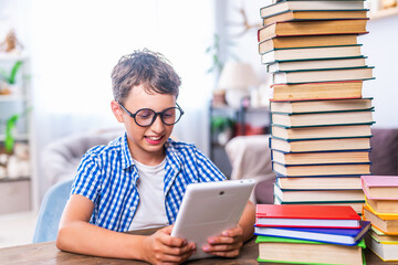 Portrait of a happy schoolboy with a tablet computer. A boy child sits at a table with stacks of books, and plays a game, uses a digital tablet for online lessons and information retrieval.