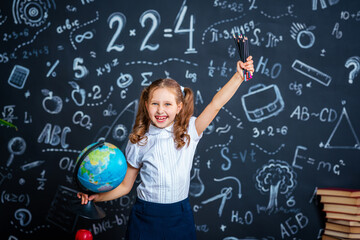 charming mischievous little girl in school uniform, smiling at camera and holding globe and pencils...