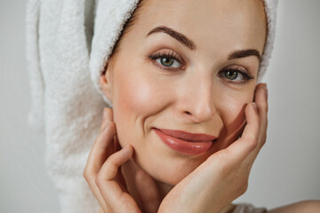 Portrait of smiling caucasian woman with healthy skin in towel posing over grey background. Happy young lady touching her perfect face with hands.