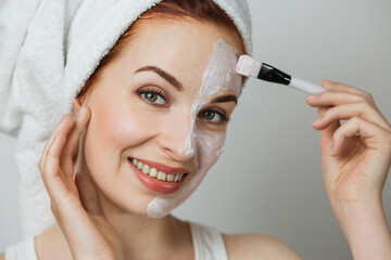 Caucasian young woman in towel using brush for applying white cosmetic cream on half of face over grey studio background. Close up, copy space.