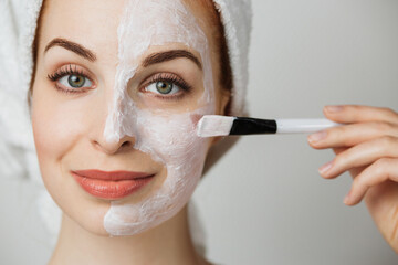 Portrait of caucasian young woman with using brush for applying facial skincare mask posing over grey background. Attractive female with professional cosmetic in studio.