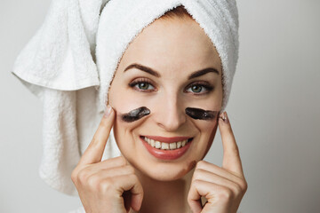 Beautiful young woman applying black face cream and looking at camera on grey studio background....
