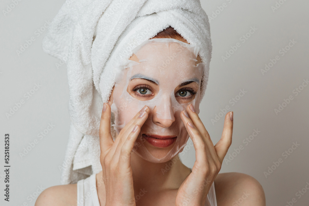 Wall mural Stunning caucasian lady in towel applying paper mask sheet on face in studio. Young woman enjoying skin care procedures over grey background.