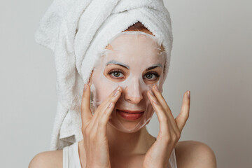 Stunning caucasian lady in towel applying paper mask sheet on face in studio. Young woman enjoying...