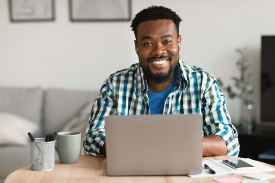 Cheerful Black Man At Laptop Computer Wearing Earbuds Working Indoors