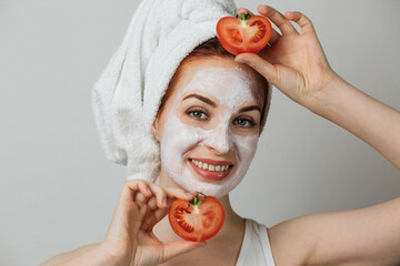 Smiling young woman with white clay mask on face holding fresh tomato against grey background....