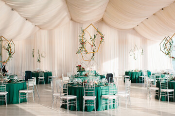 The decor of the wedding banquet hall with tables in emerald green, white drapery on the ceiling, and gold geometric decorative elements.